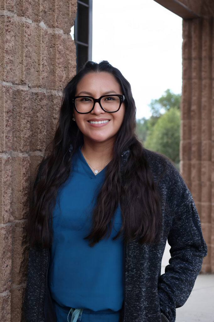 Chanda Duncan - Young female healthcare provider in blue scrubs, black sweater, and glasses smiles to camera outside